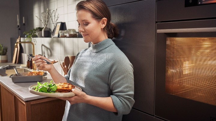 En kvinne står på kjøkkenet og spiser lasagne. 